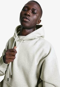 Világosszürke kapucnis pulóver, puha anyagból, laza szabású. Nyaknál húzózsinórral és bordázott mandzsettákkal rendelkezik. Részletei között szerepel egy kenguruzseb.