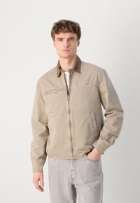 Chaqueta beige con cremallera y cuello abotonado, que presenta un bolsillo en el pecho, bolsillos laterales y una textura de tela suave. Llevada sobre jeans gris claro.