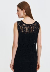 Czarny bezrękawnik z koronkowym haftem, z finezyjnymi wzorami koronkowymi, falbaniastym dekoltem i miękką teksturą. Projekt jest półprzezroczysty z tyłu.