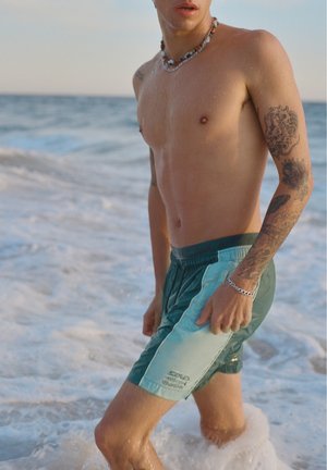 Jeune homme torse nu avec des tatouages et un collier de perles, debout dans les vagues de l'océan portant un short de bain vert, vu de la taille vers le haut.