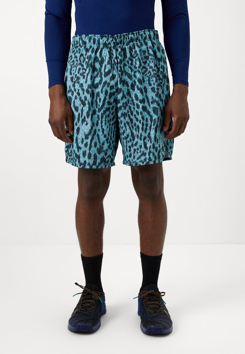 Homme portant un short bleu à imprimé léopard, un haut à manches longues bleu marine, des chaussettes noires et des chaussures de sport bleues, debout sur un fond blanc.