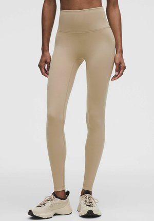 Gambe con leggings beige a vita alta e scarpe da ginnastica beige con dettagli neri su uno sfondo chiaro e semplice.