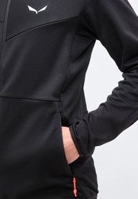 Μαύρο μπουφάν με φερμουάρ, με το χέρι ενός ατόμου στην πλευρική τσέπη με φερμουάρ, που διαθέτει μικρό λευκό λογότυπο στο στήθος και πορτοκαλί κορδόνι τράβηγμα φερμουάρ.