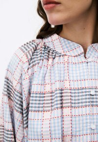 Blauwe en witte geblokte blouse met grijze accenten, rode stiksels, een staande kraag en knoopdetails aan de voorkant. Geweven stof.