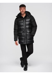 Hombre con abrigo de plumas negro con capucha, pantalones negros y zapatos negros, de pie con una mano en el bolsillo frente a un fondo liso.