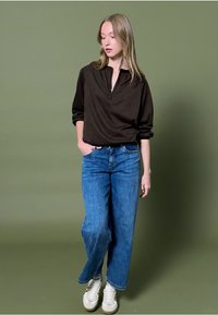 Bruine pullover met ritssluiting, losse blauwe spijkerbroek en witte sneakers. Voor een effen groene achtergrond. Vrouwelijke, toch ontspannen stijl.