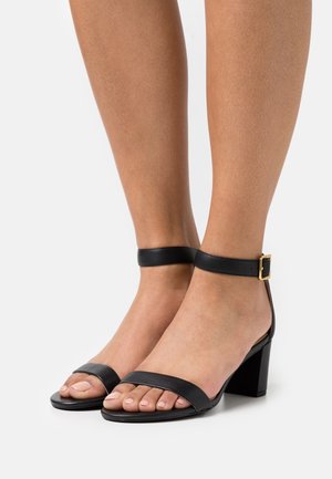 Sandales à talon carré en cuir noir avec larges brides sur le pied et une bride à la cheville ornée d'une boucle dorée. Texture lisse, design à bout ouvert.