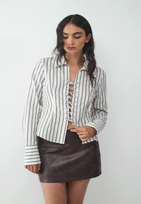 White and striped shirt with lace-up front detailing and long sleeves, paired with a brown leather mini skirt. Smooth texture, casual design.