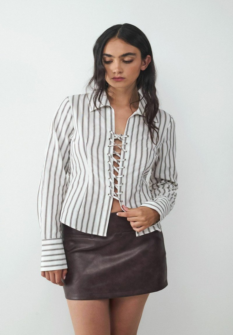 White and striped shirt with lace-up front detailing and long sleeves, paired with a brown leather mini skirt. Smooth texture, casual design.