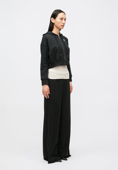 Hoodie preto curto com fecho de correr e dois bolsos frontais, sobreposto a uma camisa bege de manga comprida e combinado com calças pretas de pernas largas.