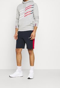 Felpa grigia con strisce rosse e blu navy, tasca frontale, abbinata a pantaloni corti blu navy con dettagli rossi, indossata con sneakers e calzini bianchi.