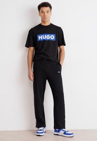 Zwarte t-shirt met blauw "HUGO" logo, gecombineerd met zwarte joggers. Model draagt blauw-witte sneakers en staat tegen een effen achtergrond.