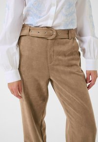 Pantalons en suède marron avec un design à jambes larges et une ceinture assortie dotée de quincaillerie en argent. Le tissu semble doux et texturé.