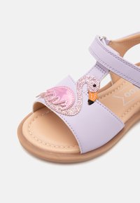 Violetti sandaali, jossa on vaaleanpunainen flamingosuunnittelu, jossa on glitterkorostuksia ja oranssi nokka. Sileä materiaali beigeillä sisäpohjilla.