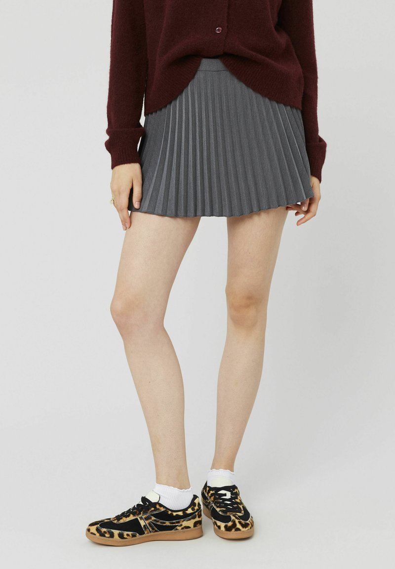 Jupe plissée grise avec une taille ajustée ; portée avec des baskets noires à imprimé léopard et des chaussettes blanches jusqu'à la cheville. Détail de tissu texturé visible.