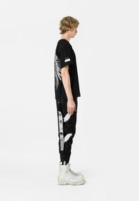 Camiseta gráfica negra con estampado de alas en la parte posterior, combinada con pantalones negros con patrones blancos y botas blancas con cordones.