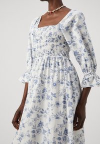 Vit klänning med blå blommönster, fyrkantig smockad liv, puffärmar och volangmuddar, med en flödande kjol och mjukt material.