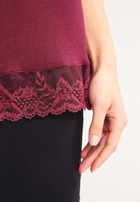Burgunderfärgad topp med spetsdetalj vid nederkanten. Spetsen har blommönster och ligger ovanpå ett svart plagg, vilket framhäver de kontrasterande texturerna.