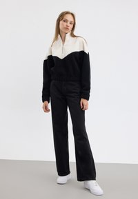 Felpa in fleece bianca e nera con design a zigzag, abbinata a jeans neri a vita alta con gamba svasata e sneakers bianche.