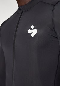 Haut de sport noir en tissu extensible, doté d'une fermeture éclair à l'avant et d'un logo blanc sur la poitrine gauche. Texture lisse avec des coutures contrastantes.