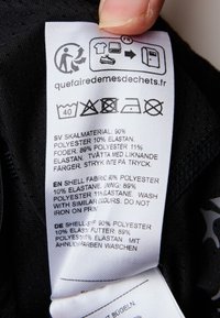 Hoitoetiketti mustassa kankaassa, joka näyttää hoito-ohjeet ja materiaali koostumuksen: 90 % polyesteriä, 10 % elastaania; sisältää useita kielikäännöksiä.