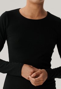 Osoba ubrana w dopasowaną czarną koszulę z długim rękawem, ręce delikatnie złożone z przodu, neutralne, jasne tło.