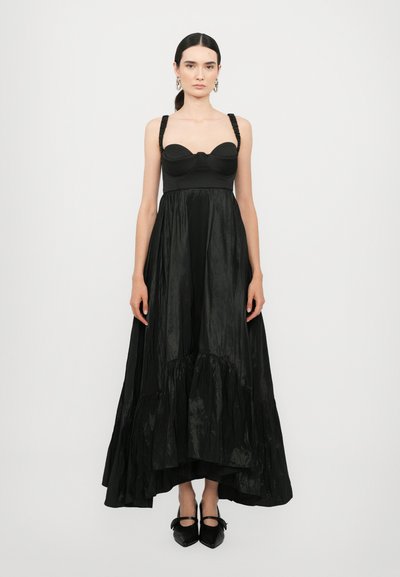 Robe noire avec un corsage ajusté et une jupe évasée texturée. Présente des bretelles tressées et un ourlet froncé. Accompagnée de chaussures noires avec des nœuds.