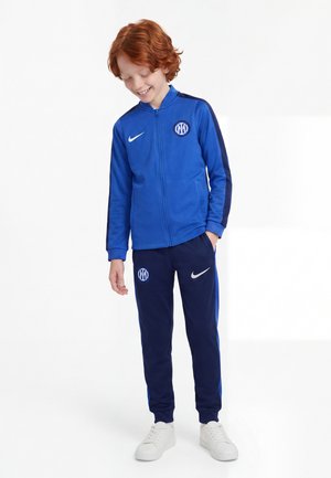 Roodharig kind glimlacht, draagt een blauwe Nike sportjas en broek met witte sneakers, staat met één hand in de zak.