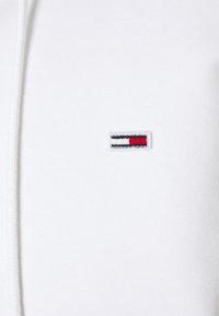 Hoodie blanco con un pequeño logo bordado y rayas horizontales rojas, negras y blancas en la parte izquierda del pecho. Material suave de algodón.