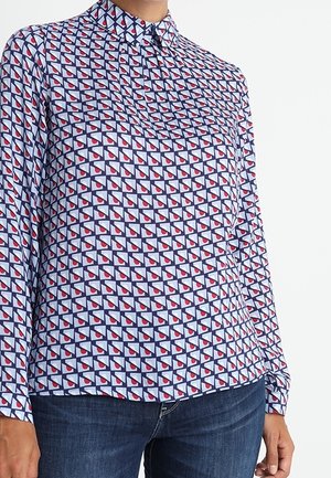 Femme portant une chemise boutonnée à manches longues avec un motif géométrique bleu, blanc et rouge, associée à un jean bleu foncé.