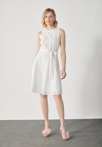 White sleeveless dress featuring eyelet pattern, A-line shape, and a tied waist. Paired with light pink shoes adorned with floral accents.