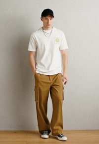 White cotton t-shirt with a green logo, paired with brown cargo pants featuring side pockets and black sneakers with white soles.