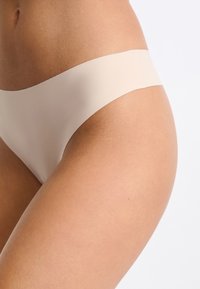 Culotte en thong beige en tissu lisse et extensible avec une taille haute minimaliste, conçue pour le confort et un port discret.