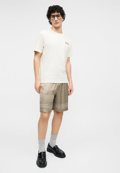 Jeune homme aux cheveux bouclés et portant des lunettes, vêtu d'un t-shirt blanc, d'un short à motifs jusqu'aux genoux, de chaussettes grises et de mocassins noirs.