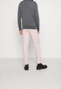 Grauer Strickpullover mit langen Ärmeln, rosa maßgeschneiderte Hose und schwarze Schuhe, von hinten gesehen vor einem schlichten weißen Hintergrund.