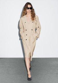 Beiger Trenchcoat mit doppelt geknöpftem Design, schwarzen Knöpfen und einem Bindegürtel, kombiniert mit dunklen Schuhen und großen schwarzen Sonnenbrillen.