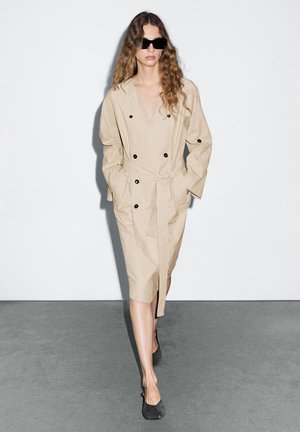 Beige trenchcoat met een dubbelgeknoopte ontwerp, zwarte knopen en een strikceintuur, gecombineerd met donkere schoenen en grote zwarte zonnebril.