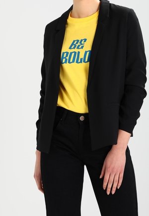 Osoba ubrana w czarną marynarkę i spodnie oraz żółtą koszulkę z niebieskim napisem „BE BOLO” na białym tle.