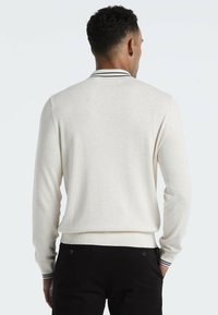 Cremfarbener Pullover mit langen Ärmeln, gerippten Bündchen und einem gestreiften Kragen. Über einer schwarzen Hose getragen, zeigt eine schlichte Rückansicht.