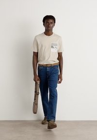 Beige bomulls t-skjorte med trykk på frontlommen, blå denim jeans og brunbelte. Bærer en lysebrun skulderveske.