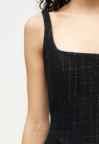 Γυναίκα που φοράει μαύρο αμάνικο φόρεμα με υφή, με λεπτές ασημί κάθετες και οριζόντιες γραμμές σε διακριτικό μοτίβο πλέγματος.