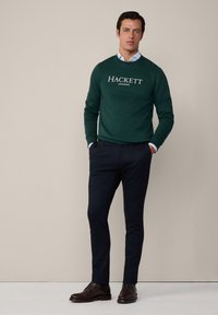Zelená mikina s potlačou "HACKETT LONDON" v bielej farbe, v kombinácii s tmavými, textúrovanými nohavicami a hnedými koženými topánkami. Model stojí na neutrálnom pozadí.