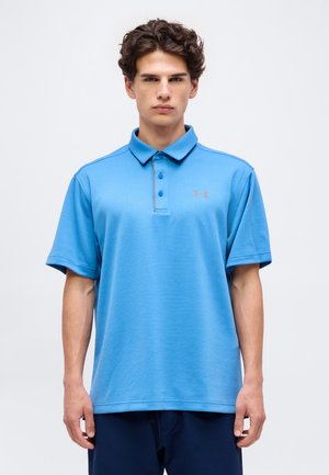 Jonge man met kort krullend haar, draagt een blauw Under Armour polohemd en donkere broek, staand voor een effen achtergrond.