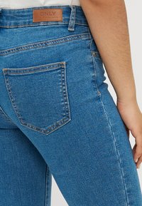 Μέτριου ύψους μπλε τζιν παντελόνι με λεία υφή, διαθέτει δερμάτινη ετικέτα στο πίσω μέρος και μία μόνο πίσω τσέπη με αντίθεση στη ραφή.