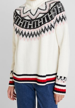 Kremowy sweter z dzianiny z czarnymi, różowymi i czerwonymi geometrycznymi wzorami, z prążkowanym dołem i mankietami, noszony z niebieskimi dżinsami, ręce swobodnie opuszczone wzdłuż ciała.