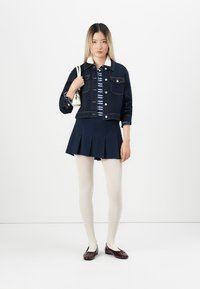 Denimjakke i mørkeblå, stripete skjorte, marineblå plissert skjørt, beige strømpebukser og brune ballettsko. Modell som holder en liten hvit håndveske.