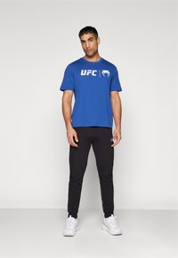 Blå kortærmet t-shirt med hvidt UFC-logo, kombineret med sorte joggers med sidelommer og ankelmanchetter, afsluttet med hvide sneakers.