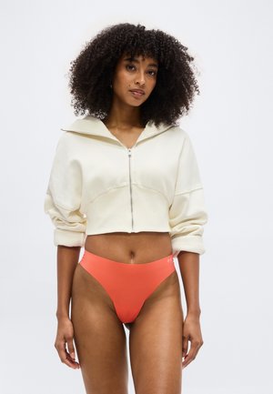 Γυναίκα με μαλλιά σε μπούκλες, φορώντας μια κομμένη λευκή φούτερ με φερμουάρ και κοραλλένιο εσώρουχο με ψηλή μέση, στέκεται μπροστά σε λευκό φόντο.