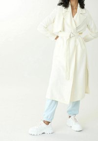 Světle béžový trenčkot s páskem v pase, širokými klopami, dlouhými rukávy a bočními kapsami, spárovaný se světle modrými tepláky a bílými teniskami.