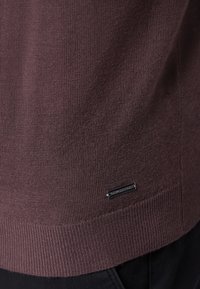 Brauner gestrickter Pullover mit geripptem Saum, der ein kleines silbernes Etikett an der Seite enthält. Die Textur ist glatt und engmaschig.